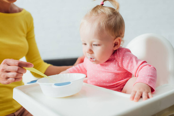 Enfoque selectivo de la niña sentada en silla alta cerca de la madre con cuchara y tazón de comida para bebés  - Foto, Imagen