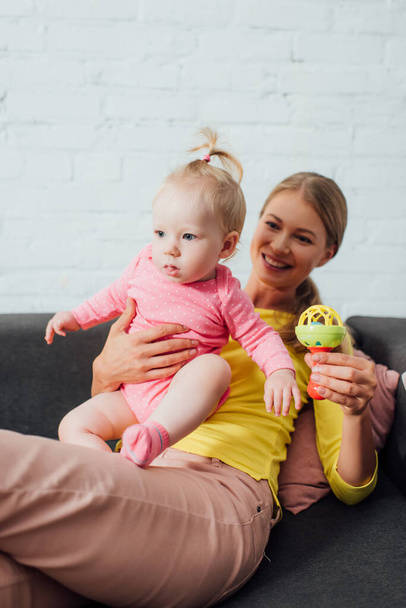 Selective focus of woman holding toy and infant daughter on couch  - Foto, Bild