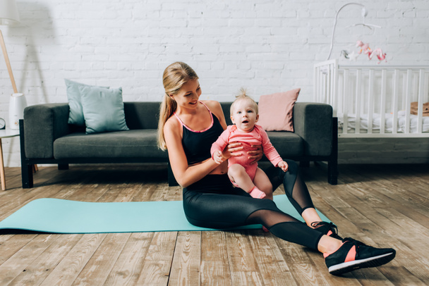 Sportswoman hugging baby girl while sitting on fitness mat at home  - Фото, зображення