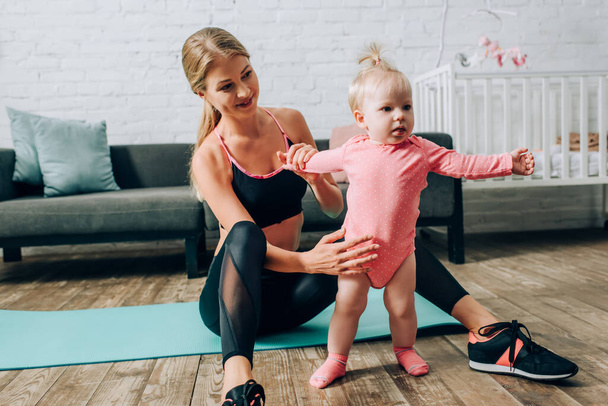 Sportswoman holding infant daughter while sitting on fitness mat in living room  - 写真・画像