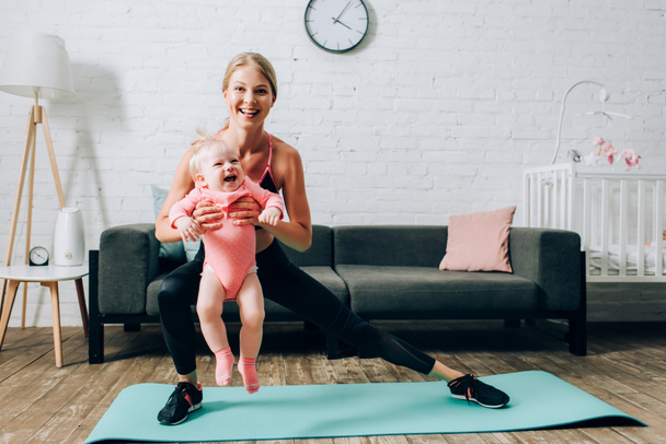 Deportiva haciendo saltos en la alfombra de fitness y llevando a su hija bebé  - Foto, Imagen