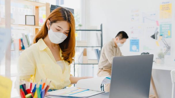 Asiatinnen tragen Gesichtsmasken zur sozialen Distanzierung in neuen Normalsituationen zur Virenprävention, während sie Laptop benutzen und durch Acryl-Trennwände getrennt im Büro stehen. Leben und Arbeit nach Coronavirus. - Foto, Bild