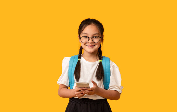 Chinees schoolmeisje met behulp van mobiele telefoon Staande over gele achtergrond, Studio - Foto, afbeelding