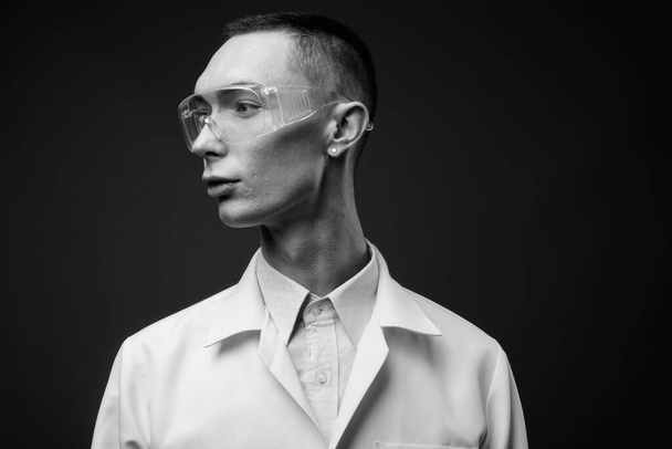 Jonge knappe androgyne man arts als wetenschapper met beschermende bril tegen grijze achtergrond in zwart-wit - Foto, afbeelding