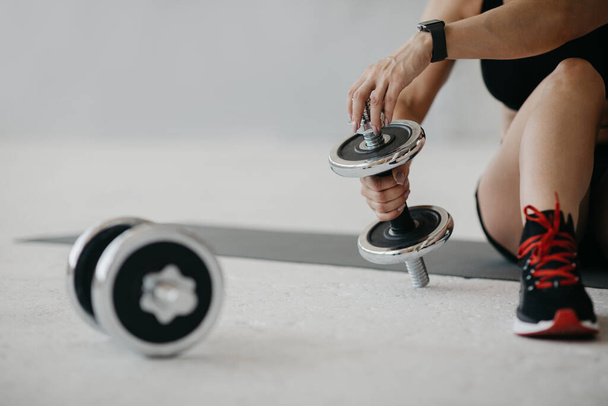 Addestramento per bicipiti. Donna adulta con smartwatch si siede sul pavimento a casa e regola il peso del manubrio per esercizi manuali - Foto, immagini