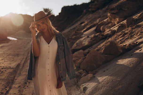 美しいブロンドの女性の帽子と白いドレスでデニムジャケットは砂漠の道を歩く - 写真・画像