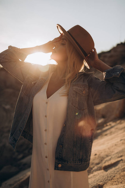 Mooie blonde vrouw in een hoed en witte jurk met een denim jas loopt op een woestijn weg - Foto, afbeelding