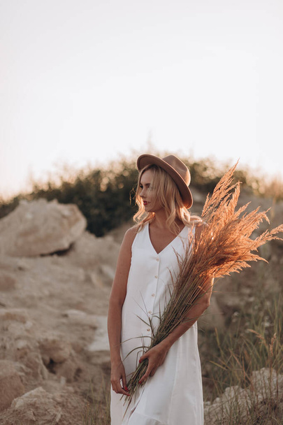 Hermosa mujer rubia en un sombrero y vestido blanco con una chaqueta de mezclilla camina por un camino desierto - Foto, Imagen