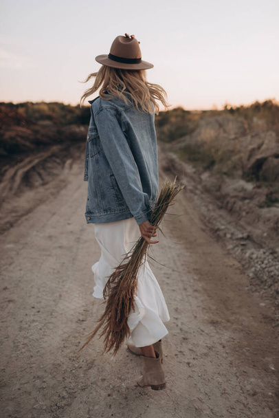 Mooie blonde vrouw in een hoed en witte jurk wandelingen op een woestijn weg - Foto, afbeelding