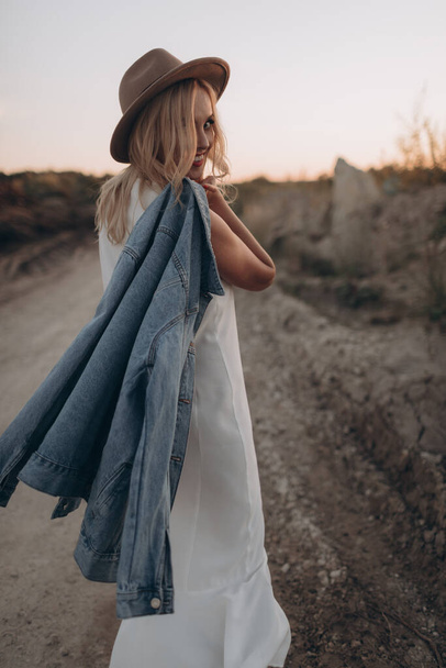Hermosa mujer rubia en un sombrero y vestido blanco camina por un camino desierto - Foto, imagen