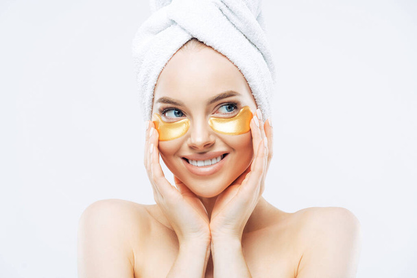 Young beautiful woman with healthy fresh skin, applies golden patches under eyes against dark circles, shows effect of perfect cosmetic product, stands shirtless, isolated on white background. - Φωτογραφία, εικόνα