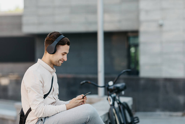 Oblíbená hudba, kolo a město. Usmívající se chlap s moderními sluchátky, dívající se na smartphone na ulici - Fotografie, Obrázek