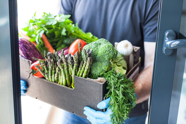 Fresh organic greens and vegetables delivery during coronavirus Covid-19 pandemic outbreak. Man hands wearing blue latex medical gloves holding box with safe grocery delivering in the house doorway - Valokuva, kuva
