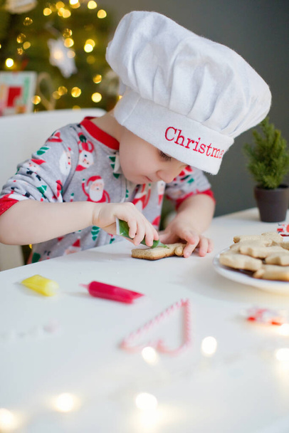 Söpö lapsi poika koristaa joulun evästeet värikkäillä kuorrutteilla valkoisella pöydällä lähellä joulukuusta valoilla. Joulun ruoanlaitto käsite - Valokuva, kuva
