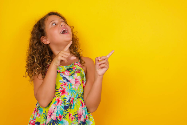 Advertisement concept. Positive little girl wearing summer dress on yellow background  pointing her two index finger and looking on empty copy space. - Foto, Imagen