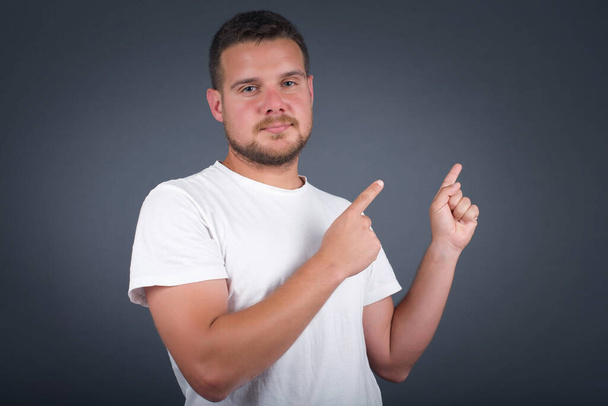 Pretty young man with positive expression, dressed casually, indicates with  fingers at blank copy space for your promotional text or advertisement. Adorable man poses indoor alone - Photo, Image