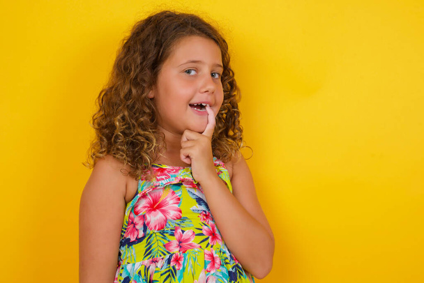 Carefree successful little girl wearing summer dress on yellow background entrepreneur giving lecture how become confident businesswoman smiling broadly self-assured touching jawline gazing camera tilting head grinning white teeth delighted - Photo, Image