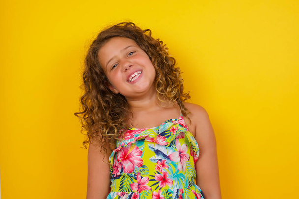 mooi klein meisje draagt zomer jurk op gele achtergrond met brede glimlach, toont witte tanden, draagt casual t-shirt, staat over grijze studio muur vreugde met een dag vrij. vrouw die binnen staat. - Foto, afbeelding