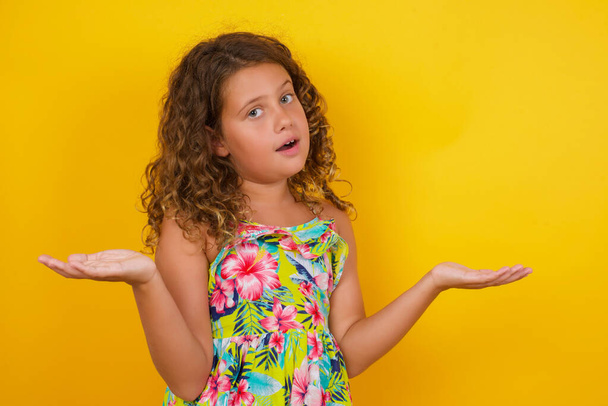 So what? Portrait of arrogant unbothered good-looking modern young stylish little girl wearing summer dress on yellow background, shrugging careless hands sideways smiling gasping indifferent, telling something obvious. - Photo, Image