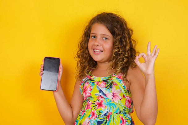 Захоплена маленька дівчинка в літній сукні на жовтому тлі, що показує порожній екран смартфона, миготливий погляд і робить знак з рукою. Студійний знімок шокованої дівчини, що тримає смартфон з порожнім екраном. Концепція реклами. - Фото, зображення