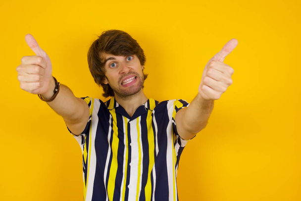 Молодчина! Портрет счастливого улыбающегося голубоглазого молодого успешного человека, подающего два больших пальца вверх, стоя в помещении. Положительная эмоция человека выражение лица язык тела.  - Фото, изображение
