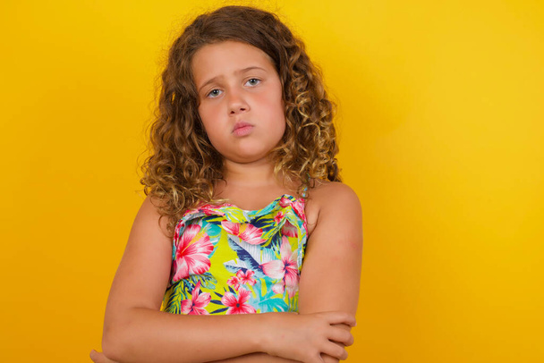 Tailliertes Porträt eines schönen kleinen Mädchens im Sommerkleid auf gelbem Hintergrund, das vor Missfallen die Stirn runzelt, lässig gekleidet ist und die Arme verschränkt hält - Foto, Bild