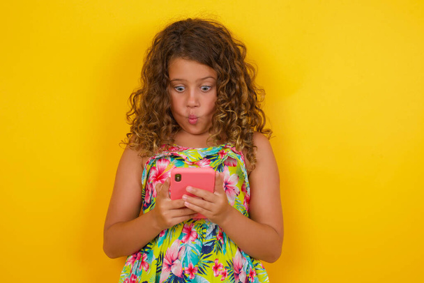 Poziome ujęcie ładnej młodej dziewczynki w letniej sukience na żółtym tle wygląda z błędnymi oczami, posiada nowoczesny smartfon, otrzymuje niespodziewaną wiadomość od znajomego, czyta przypomnienie.  - Zdjęcie, obraz