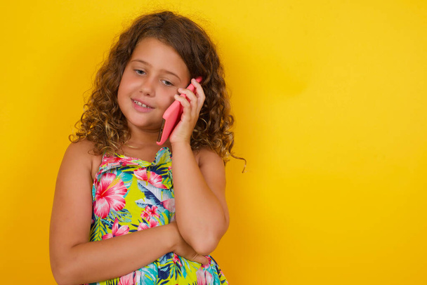 Porträt eines lächelnden kleinen Mädchens im Sommerkleid auf gelbem Hintergrund, das auf einem Mobiltelefon spricht - Foto, Bild