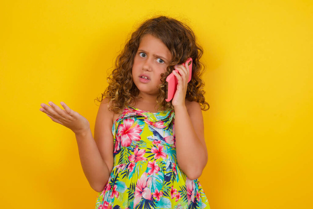 Ein kleines Mädchen im Sommerkleid auf gelbem Hintergrund, gestresst mit der Hand auf dem Gesicht, schockiert vor Scham und Überraschung, wütend und frustriert. Angst und Verärgerung über Fehler. - Foto, Bild