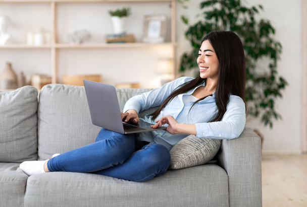 Pasatiempo favorito. Mujer asiática alegre que relaja con el ordenador portátil en el sofá, hojeando Internet - Foto, Imagen