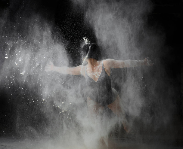 Όμορφη λευκή γυναίκα με μαύρο κοστούμι και αθλητική φιγούρα χορεύει σε λευκό σύννεφο αλεύρου σε μαύρο φόντο. - Φωτογραφία, εικόνα