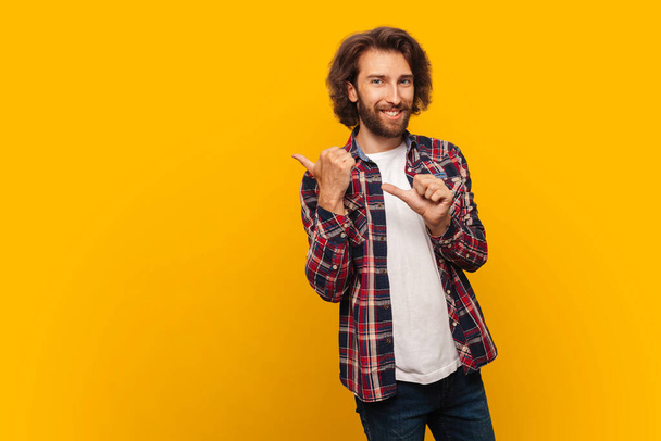 Handsome curly haired man with long hair in casual shirt standing against isolated yellow background pointing to back from behind with hand and thumbs up, smiling confidently - Photo, Image