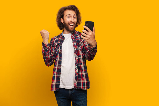 A young man with a beard rejoices holding a mobile phone in his hands. Isolated on yellow background - Фото, зображення