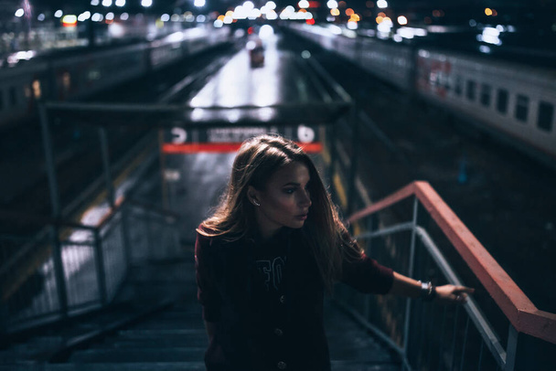 beautiful young lonely girl stands up the stairs. behind the back of the railway station the lights are reflected in puddles on the street night, it rains, she waits for the train looks to the side - Valokuva, kuva