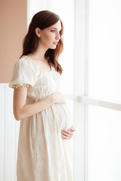 красива вагітна жінка в білій сукні, що стоїть біля вікна
 - Фото, зображення