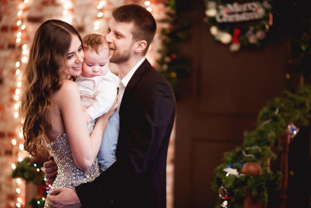 feliz familia joven en vestido y traje con su pequeño hijo en los brazos abraza, sonriendo, se queda en un hermoso interior de Navidad con una escalera de madera decorada contra una pared de ladrillo con luces - Foto, Imagen
