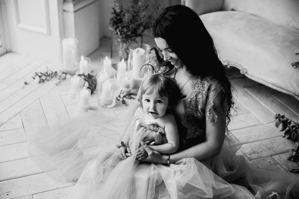 bela morena mulher mãe senta-se em um piso branco com sua filha amada em seus braços e sorri em um quarto brilhante com uma decoração de flores e velas - Foto, Imagem