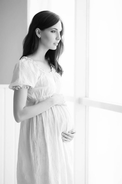 gyönyörű terhes nő fehér ruhában áll az ablak mellett - Fotó, kép