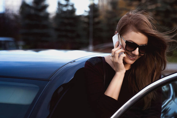 beautiful young woman in black jacket and sunglasses talking by smartphone near car - Zdjęcie, obraz