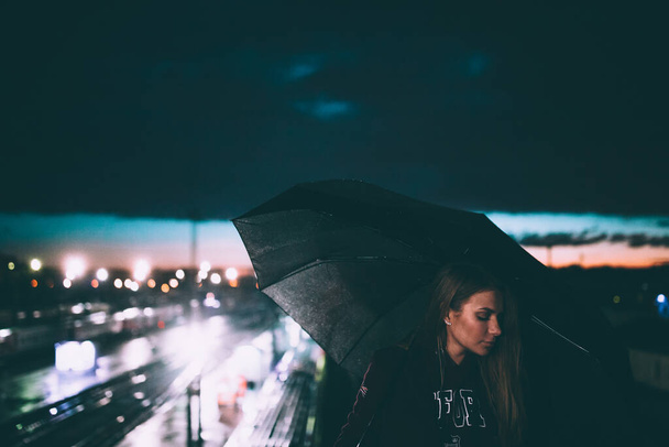 beautiful young lonely blonde girl standing near road under an umbrella on night street - Foto, Imagem