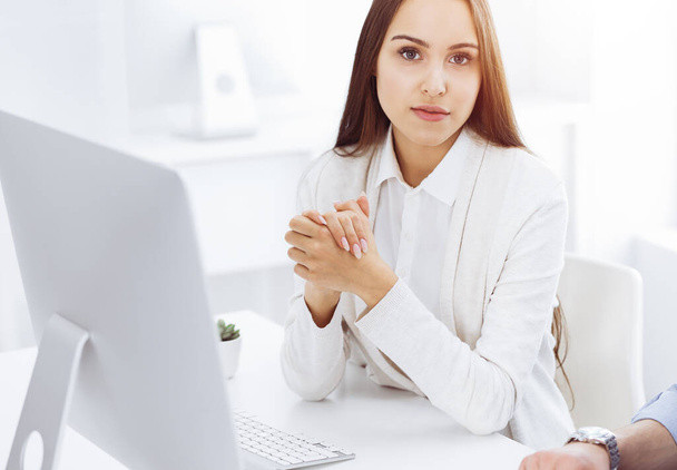 Young cheerful woman sitting at the desk with computer and looking at camera in sunny office. Looks like student girl or business lady. Headshot - Photo, Image