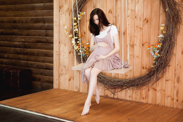pregnant woman in pink dress sitting on a swing on a wooden background - Photo, Image