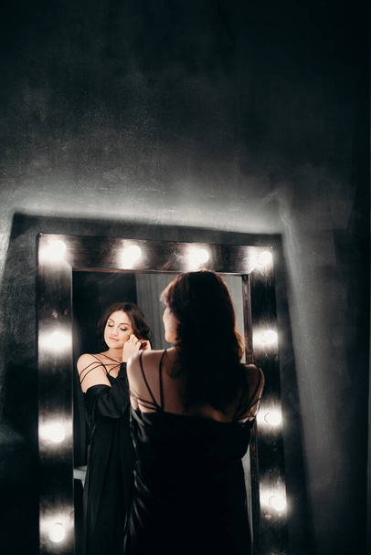 Beautiful woman stay near big mirror with lamp bulbs in dark room - Φωτογραφία, εικόνα