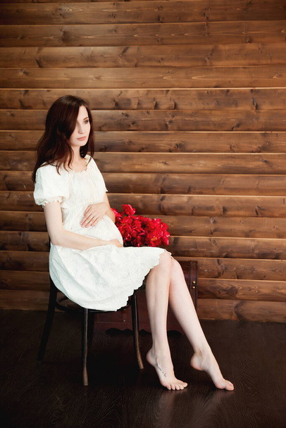 красивая беременная женщина в белом платье и цветы, сидя на стуле, деревянный фон - Фото, изображение