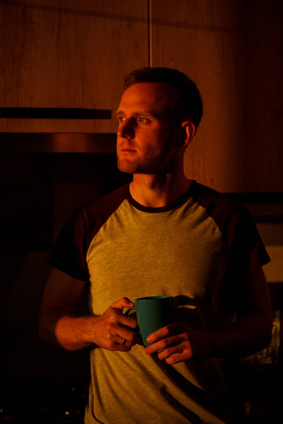A man standing in front of a window in sunset lighting is drinking coffee in his kitchen - Photo, Image