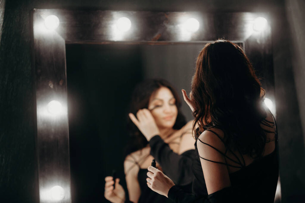 Beautiful woman makes up near big mirror with lamp bulbs. Dark background. - Photo, Image
