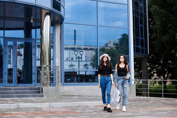 Two young brunette girls, wearing casual jeans attire, walking in front of blue modern glass building. Girlfriends traveling in city, holding backpacks, sightseeing. Active lifestyle concept. - Foto, imagen