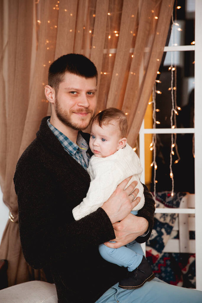 šťastný otec se synem v náručí na pozadí okna s průhlednou oponou, světly a vánočním stromečkem - Fotografie, Obrázek
