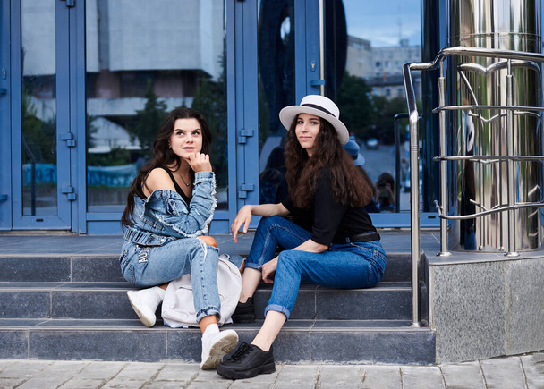 Two young brunette girls, wearing casual jeans attire, sitting on stairs of blue modern glass building. Girlfriends traveling in city, resting after walk. Active lifestyle concept. - Фото, зображення