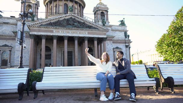 Couple is photographed on background of temple. Media. Young woman and man take selfie on bench at temple. Couple takes funny selfies on background of beautiful temple - Photo, Image